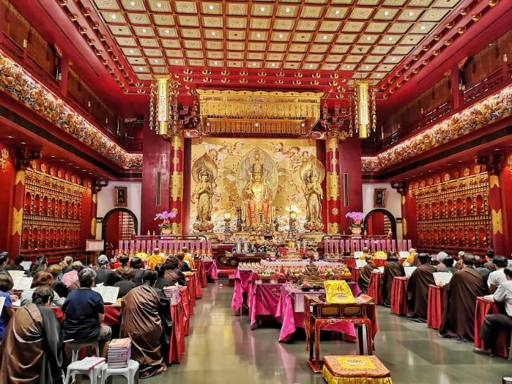 Devotees in a temple with golden Buddha statues, ornate decor, and rows of seating for prayer