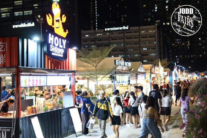 Nighttime scene at Jodd Fairs DanNeramit in Bangkok, with crowds of people walking among brightly lit food stalls