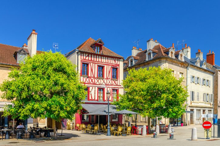 Sunlit European street with a red-and-white timber-framed building, outdoor cafe tables, and green trees under a blue sky.