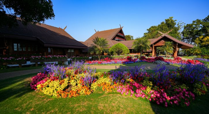 Colorful flower beds border a manicured lawn in front of rustic wooden buildings with thatched roofs and palm trees.