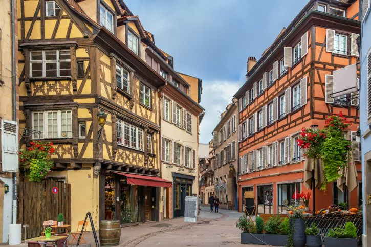 Colorful European street with timber-framed houses, outdoor cafe seating, and flower pots.