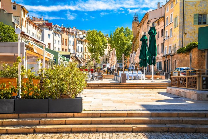 Sunny plaza with outdoor cafe seating, green umbrellas, and stone-paved square among colorful buildings.