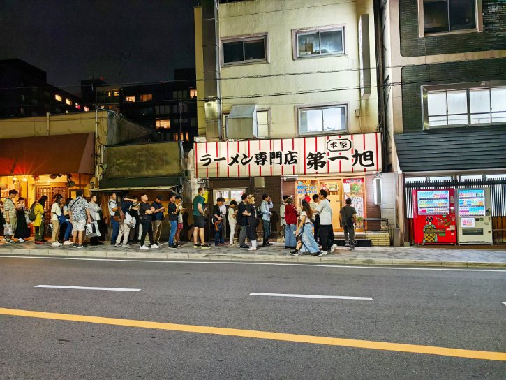 Line of people waiting outside a ramen shop at night along a city street.