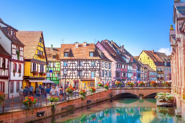 Colorful half-timbered houses line a canal with a stone arched bridge and pedestrians strolling under a blue sky.
