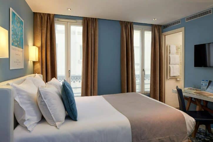 Bright hotel room with a large bed, white linens, blue accent pillows, and brown curtains by tall windows.