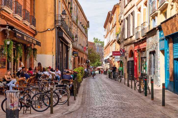 Cobbled street with outdoor cafe seating on the left and colorful historic buildings on both sides.