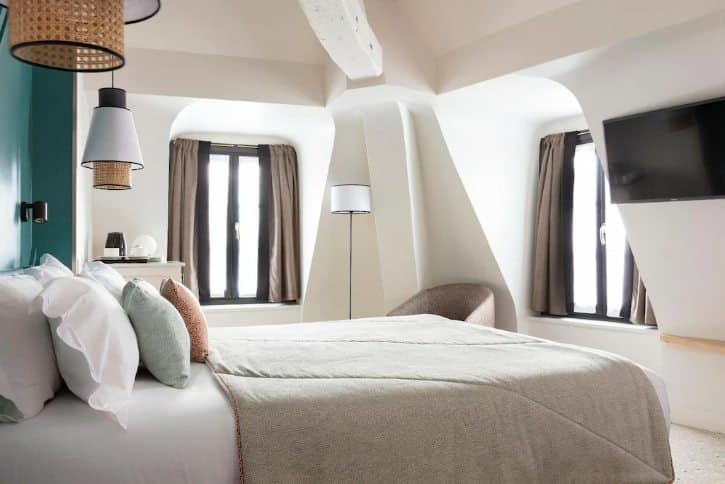 Bright attic hotel bedroom with a large bed, white linens, beige throw, and multiple pillows under sloped ceilings with dark-curtained windows nearby.