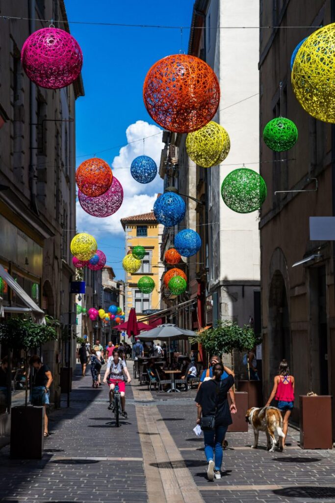 Narrow street decorated with large colorful woven spheres hanging above pedestrians and cyclists.