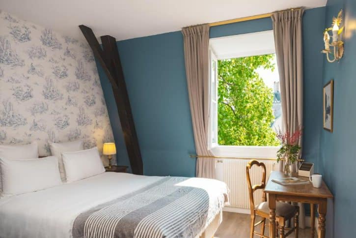 Bright hotel bedroom with a made bed, blue walls, exposed dark wooden beams, and a window with beige curtains showing green trees outside.