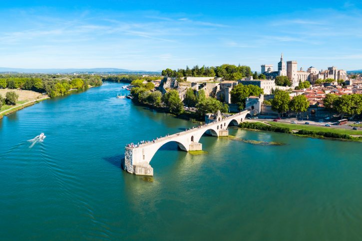 Aerial view of a stone-arched bridge crossing a wide blue river, with green riverbanks and a historic town in the background.