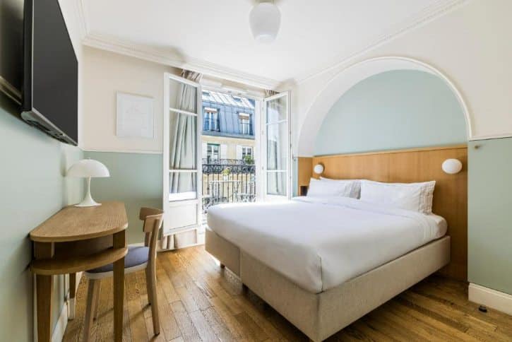 Hotel room with a double bed, open balcony doors, desk and chair, and warm wooden floors at daylight.