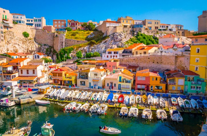Colorful hillside town by a calm harbor with boats moored along a quay and pastel houses above.