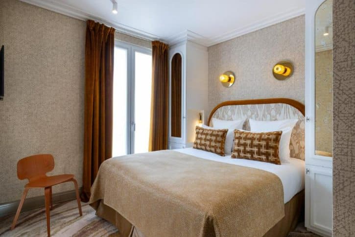 Hotel room with a double bed, beige bedding and brown patterned pillows, wooden headboard, warm wall lights, and tall windows with brown curtains.
