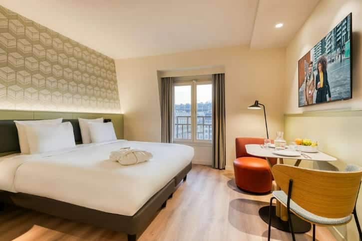Hotel room with a large bed, patterned accent wall, and a seating area by a window.