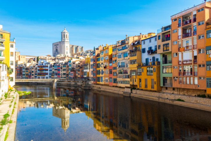 Row of colorful residential buildings lining a river canal, their reflections shimmering in the water, with a white church tower in the background.