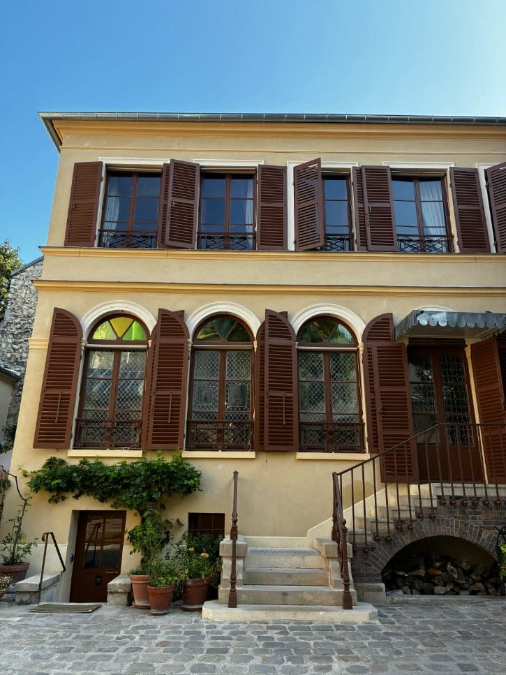 Two-story beige building with brown wooden shutters and arched windows, stone stairs, potted plants, and a cobblestone courtyard.