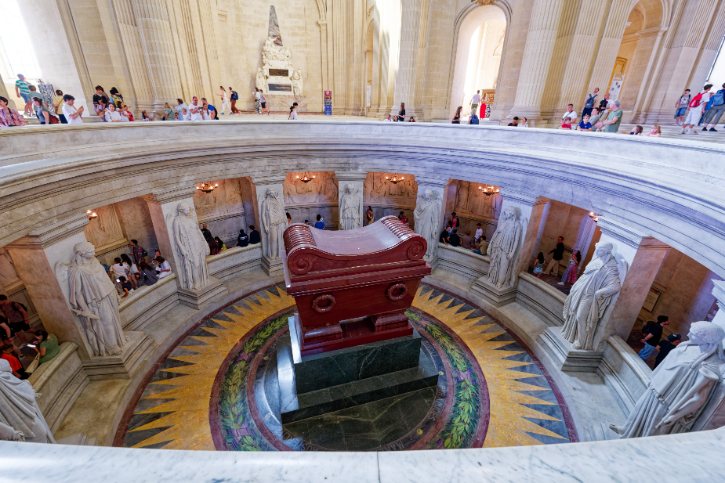 Circular rotunda with a central red sarcophagus, marble statues around, sunburst floor pattern, and visitors on the upper level.