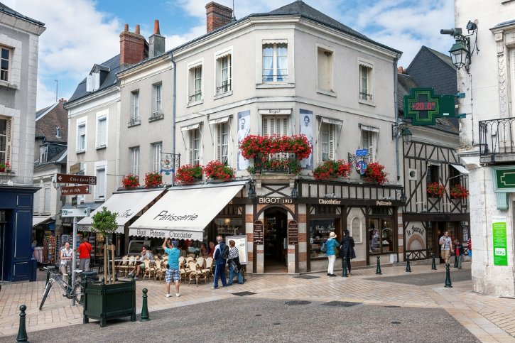 Corner street with a white three-story building housing a pâtisserie and chocolatier; red flower boxes, outdoor seating, and pedestrians.