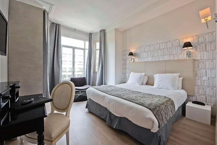 Bright hotel room with a large bed, white linens and a gray bed runner, plus a window seating nook by sheer curtains.