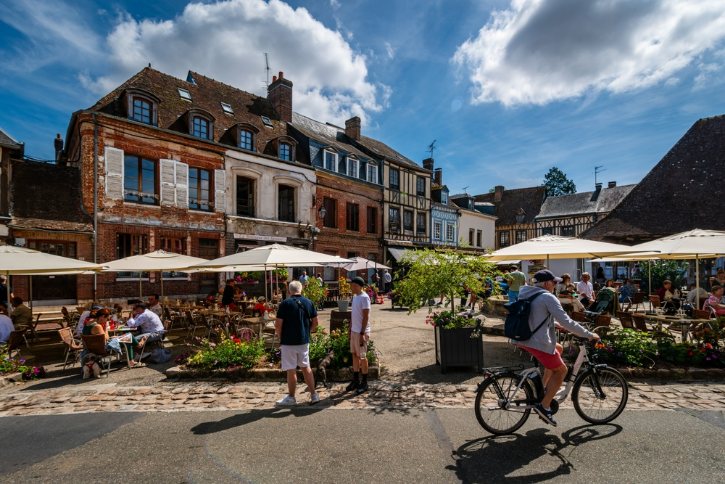 Outdoor cafe in a sunny town square with white umbrellas, diners at tables, and historic brick buildings in the background in a European village.