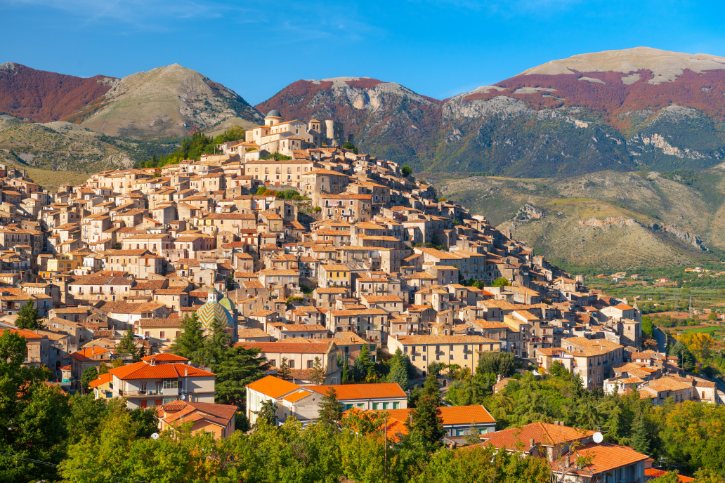 Stone hillside town with terracotta roofs perched on a slope, set against mountains in the distance.